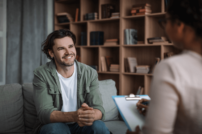 Millennial black woman psychologist consults smiling european guy in modern clinic interior. Solving of psychological problems, mental therapy and treatment, professional medical help and support