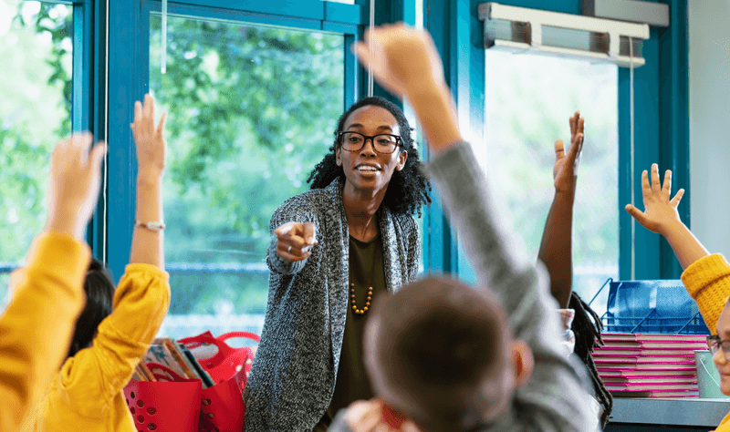 Teacher in classroom points to student raising hand - stock photo
A multiracial group of elementary students in class raising their hands to answer a question. The focus is on their teacher, an African-American and Hispanic woman in her 30s, who is standing in front, facing them, smiling and pointing to a student. Most of the students are 9 years old.